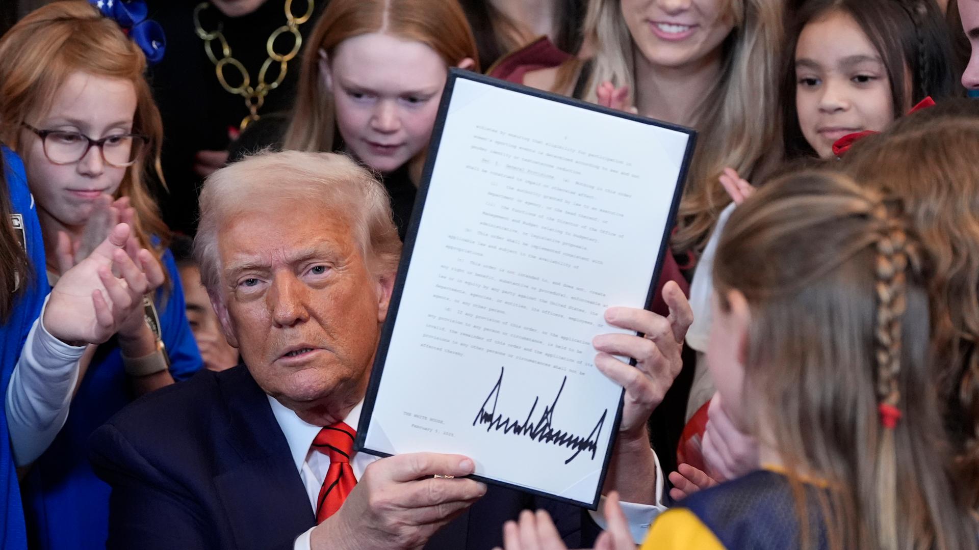 US President Donald Trump signs a implementing regulation in the White House that prohibits transsexual athletes from participating in women's and girls sports events. There are many young girls and women behind him.