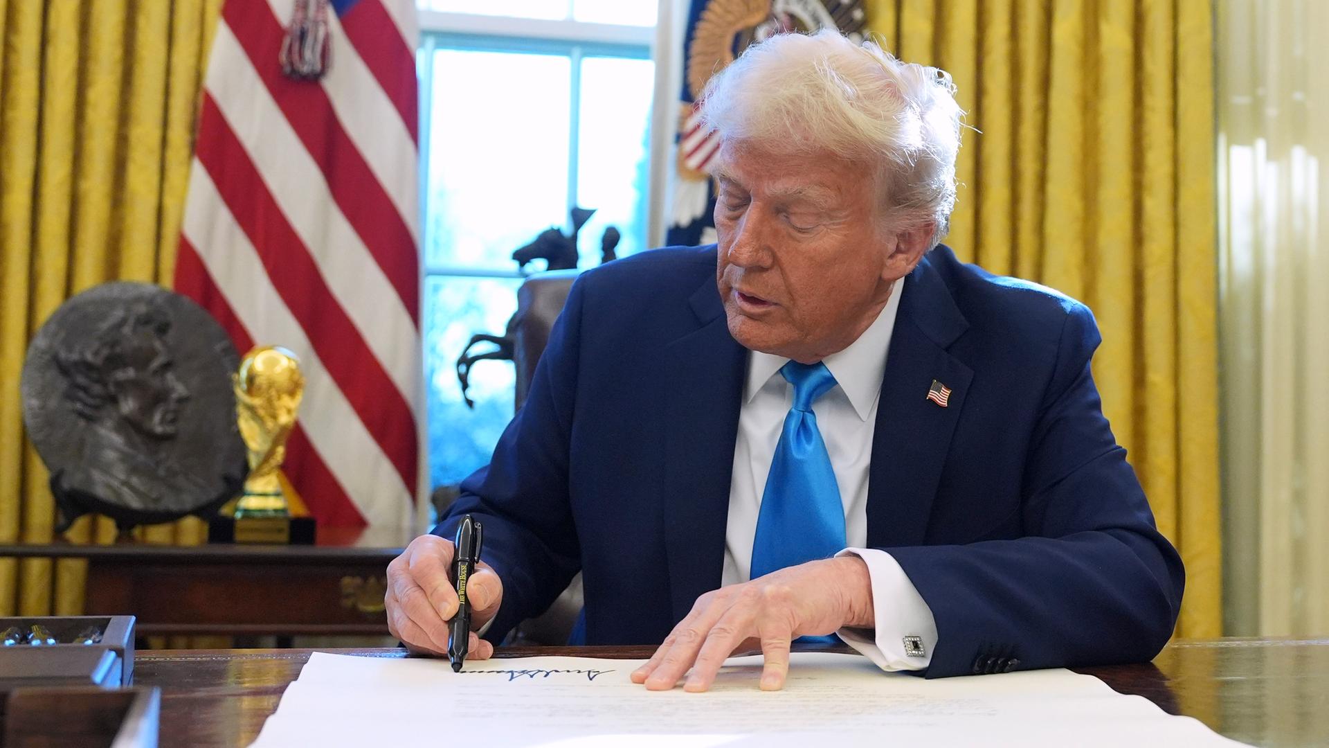 Donald Trump sits at a table in the White House and signs a document.