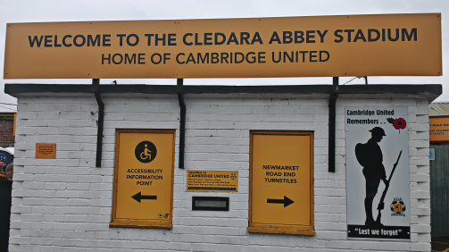 © Lennart Fey - The entrance gate to the Cledara Abbey Road Stadium of Cambridge United. On it stands on it "Welcome to the Cledara Abbey Road Stadium, Home of Cambridge United". Among them is a sign that shows to the left for admission information for people with restricted movement ability (a pictogram of a person in a wheelchair)