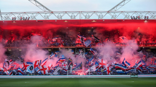 © Imago - The Fankurve Sampdorias in the Marassi Stadium on the penultimate matchday of series B 2024/25 © Imago - The picture shows the full fan curve of the Italian football club Sampdoria Genoa in a stadium. The view goes from the middle of the field directly to the stands behind the gate. The grandstand is densely filled with fans, which swirl numerous large and small flags in the club colors blue, white, red and black. Many flags show club logos, lettering or pictures of famous players. Red Bengalos and smoke torches glow in the upper area of the stands, which immerses the scene in an intense red light. In the lower area, thick smoke can be seen in pink and red tones that spreads over the fans. The atmosphere looks very loud, energetic and emotionally - typical of a so -called "Ultras"-Fank curve in an important game. The empty field can be seen in the foreground, with a goal in the middle. At the bottom right there is an electronic scoreboard that the time "00:00" shows. The stadium construction with roof and lighting can also be seen. The image scene conveys strong passion and support of the fans for their team.