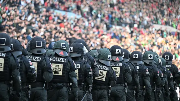 Police officers stand one behind the other and look at an occupied stadium stand.