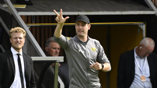 © imago - The photo shows the dugout of a football stadium during a game. There are two men in the foreground. On the left there is a man in a black suit with a white shirt and black tie: coach Oscar Hiljemark from Elfsborg. He has curly, blonde hair and looks forward with concentration. To the right stands a man in Mjällby AIF's sportswear, coach Karl Marius Aksum - gray top with club logo, black tracksuit bottoms and black cap. He raises his right arm and shows a gesture with his hand that is reminiscent of a tactical instruction. Other people can be seen in the background, including an older man with gray hair and glasses who is looking at his cell phone. The scene appears to take place during a football match, and the mood appears concentrated and tense.