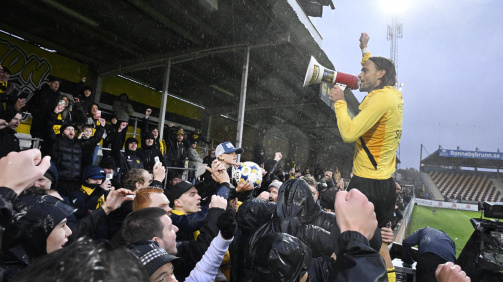 © imago - The photo shows a cheering crowd in a football stadium. In the foreground is a player from the Mjällby AIF club wearing a yellow jersey with black details. He stands elevated on the boards and holds a megaphone in his hand, which he uses to speak or sing to the fans. Many spectators in the background wear club colors of yellow and black and cheer with raised fists. It is raining lightly and some fans are wearing rain jackets or hoods. The atmosphere is exuberant and emotional – it seems like a celebration after an important victory. A covered grandstand can be seen in the background.