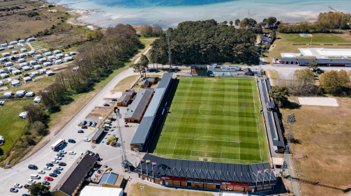 © imago - The picture shows an aerial view of the Strandvallen football stadium, the home ground of the Swedish club Mjällby AIF. The stadium is located directly on the Baltic Sea coast, with the sea and a light, sandy beach in the background. The pitch's green grass is well-maintained and has a rectangular shape surrounded by stands. The main stand is located in the lower part of the picture and is covered with a black roof. Flags and advertising signs can be seen on the roof. Next to the stadium there are parking lots with several cars, some buildings and a campsite with many white caravans. In the background there are forests and the calm, blue Baltic Sea. The atmosphere appears calm and sunny.