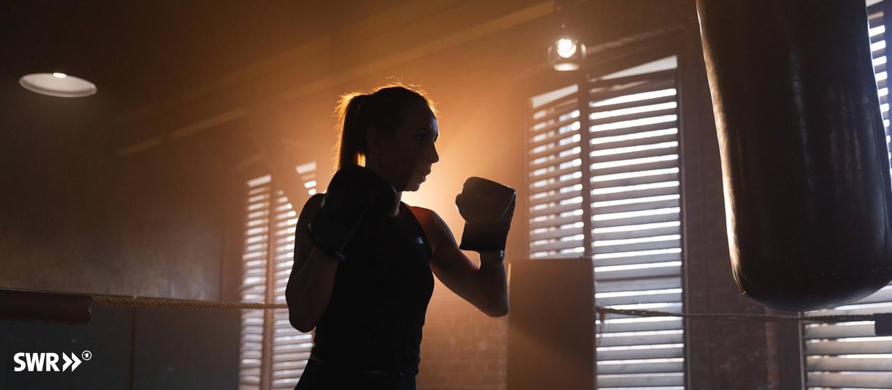 Female boxer stands in front of a punching bag, the sun shines from behind into the dark room