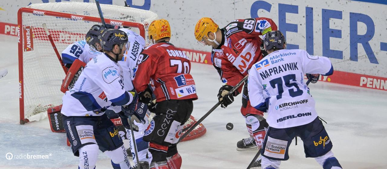 Players from the Fischtown Pinguins and the Iserlohn Roosters stand around an ice hockey goal and fight for the ball