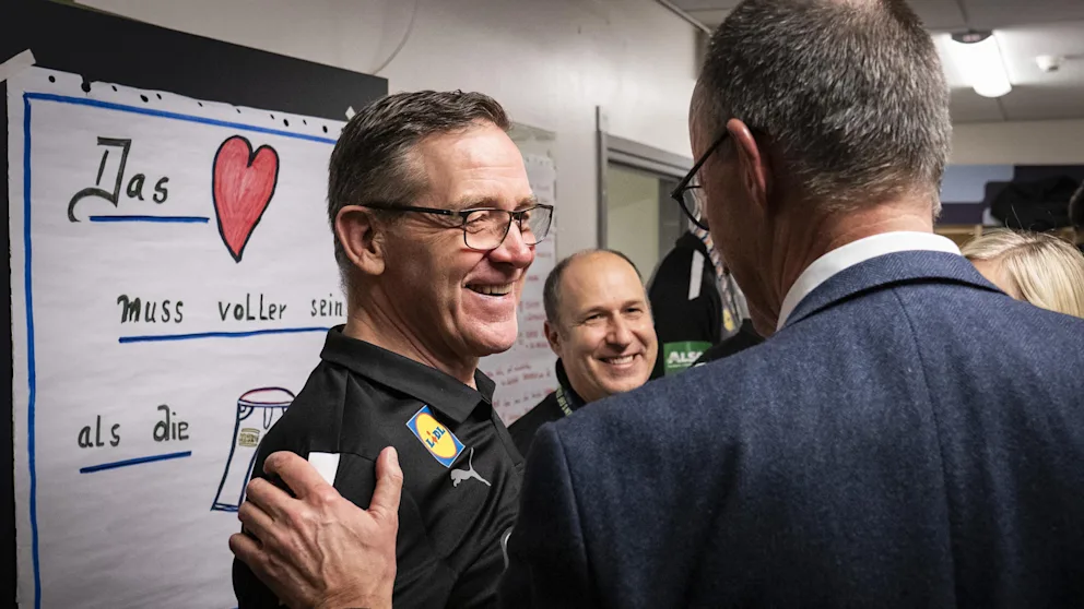 Die Chefs unter sich! Merz im Gespräch mit Bundestrainer Alfred Gíslason (66). Links zu sehen: das Kabinen-Plakat der DHB-Jungs mit dem Motto „Das Herz muss voller sein als die Hose“