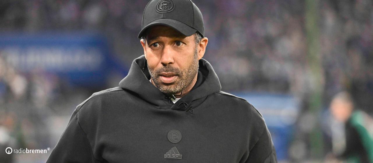 Fortuna Düsseldorf's football coach, Daniel Thioune, stands on the sidelines of the Hamburg stadium wearing a black hoodie and black cap.