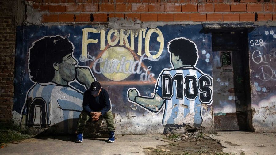 Photogallery A man sits in front of a mural depicting the late soccer star Diego Maradona and reads in Spanish Fiorito, city of God in the courtyard of the late soccer player's childhood home