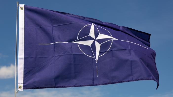 A NATO flag waving in the wind against a blue sky.
